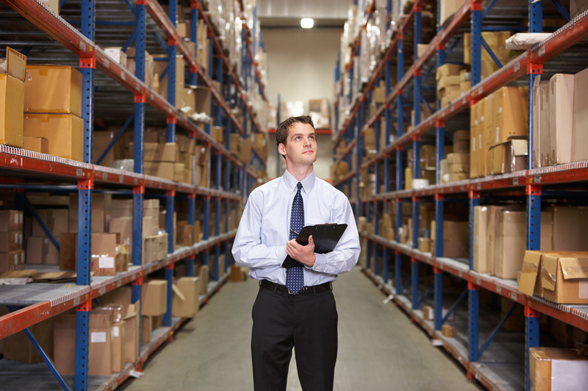 Manager In Warehouse With Clipboard Looking Up At Boxes
